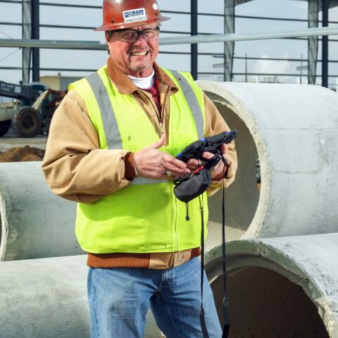 a Crain Construction employee smiling while holding an iPad on a construction site wearing a hard hat and safety vest