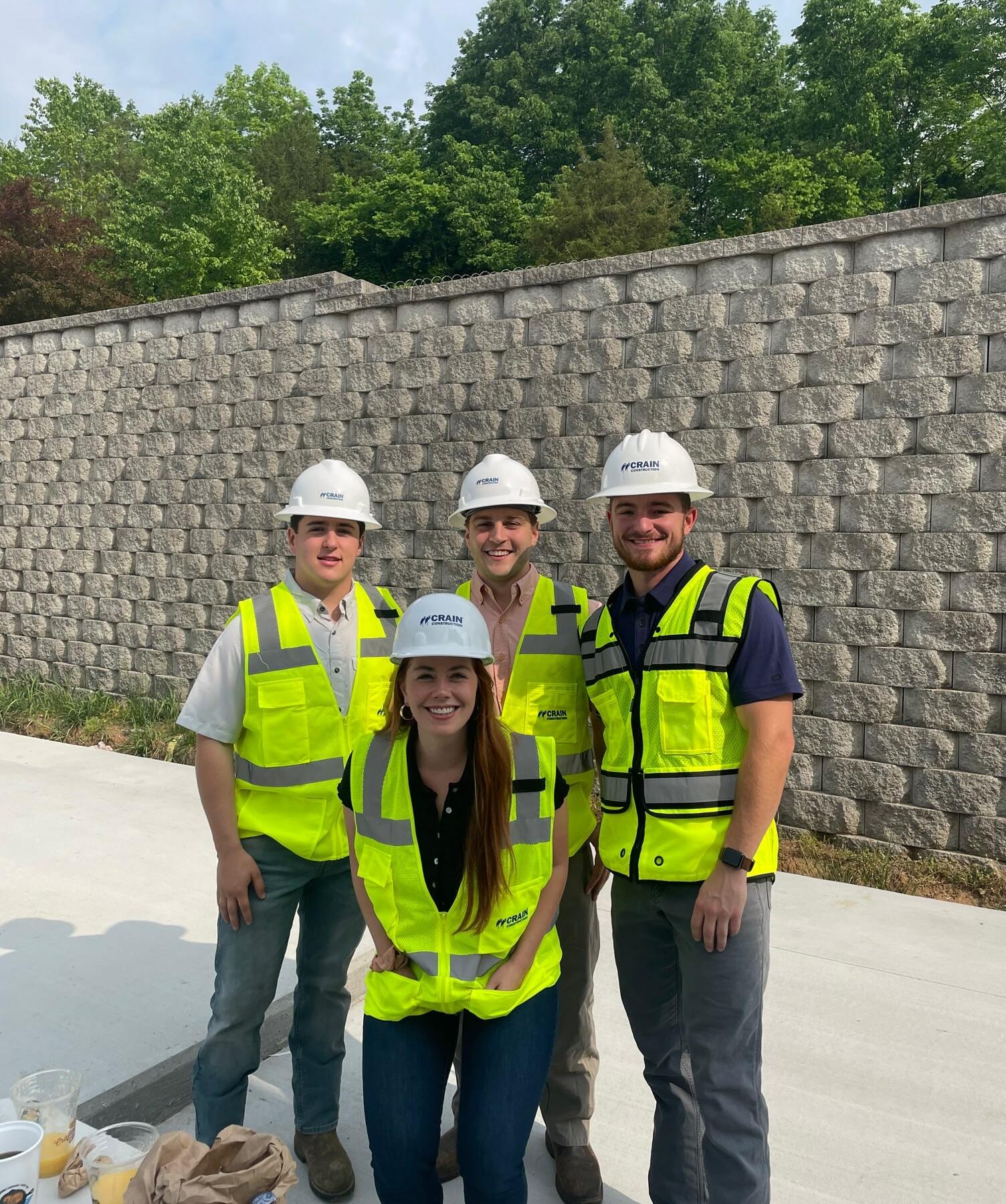4 Crain Construction workers smiling in hard hats with neon vests on in front of a brick wall